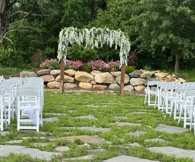 White chairs and a wisteria-draped arbor set for a ceremony beside the rock wall
